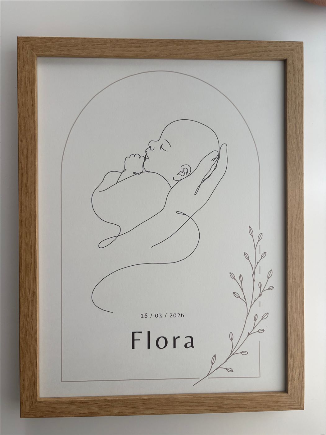 Le dessin au trait, en gros plan : un nouveau-né endormi, tenu d’une main, avec le prénom Flora et sa date de naissance.